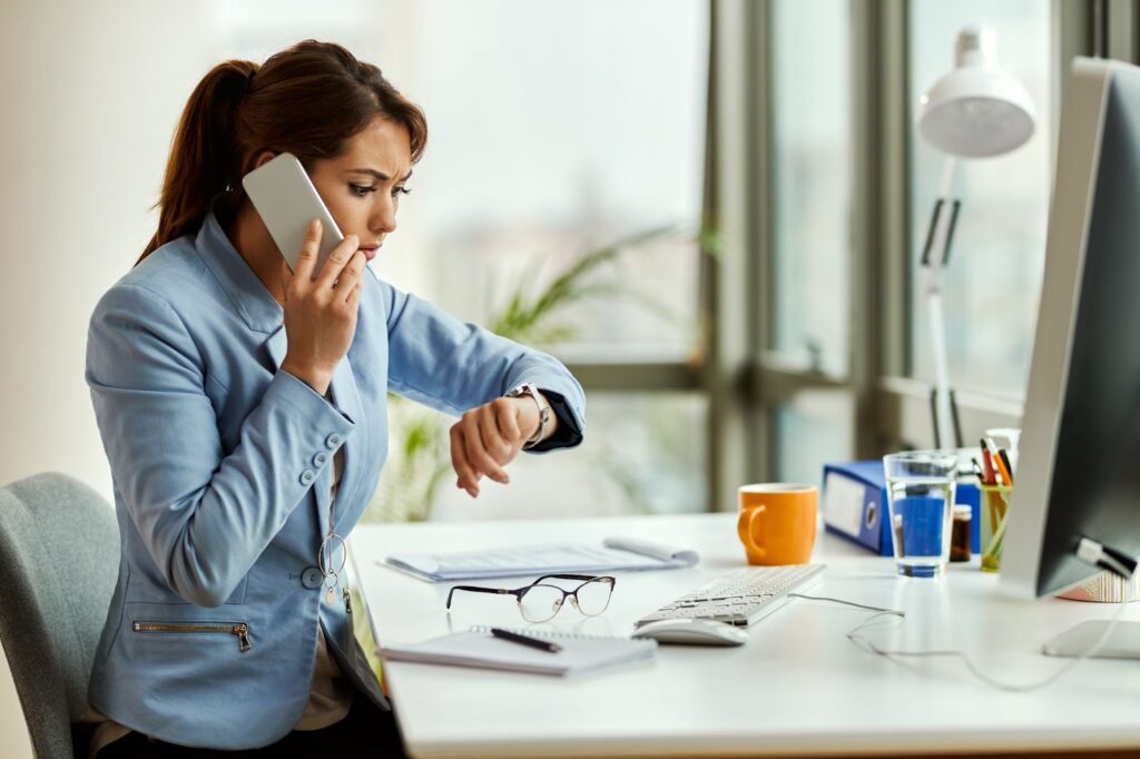 Frustrated businesswoman on a phone call, glancing at her watch while sitting at her desk.